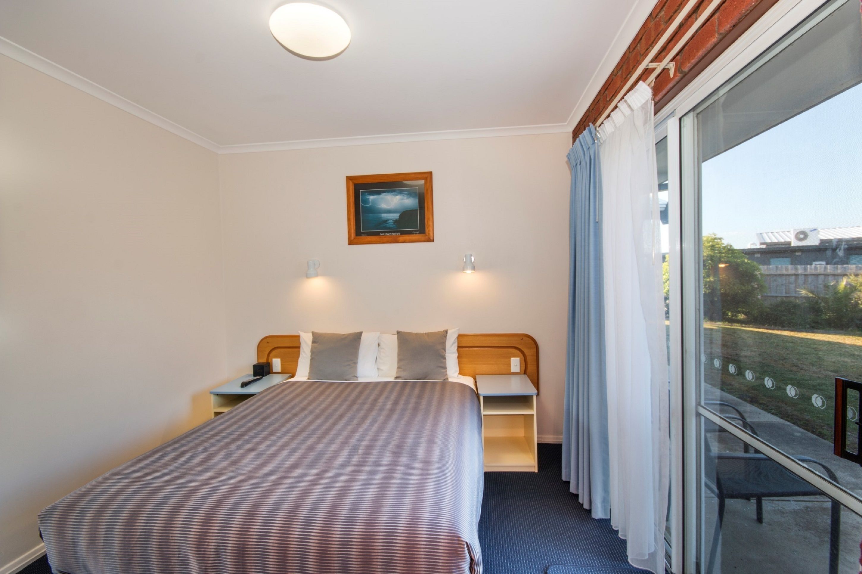 Une chambre d’hôtel avec un lit, des tables de chevet, une commode, une fenêtre avec des rideaux et un tableau encadré au mur.