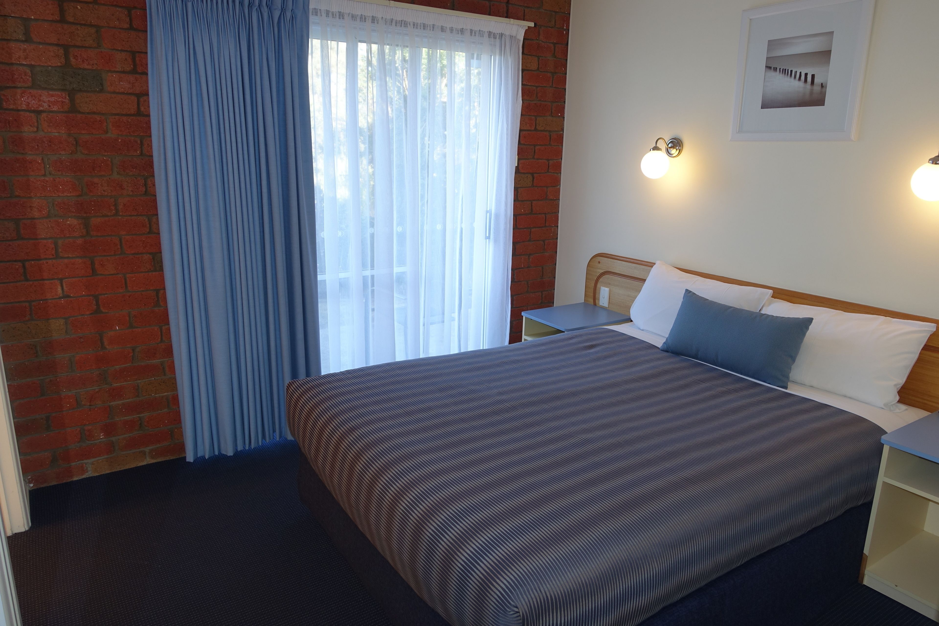 Een hotelkamer met een bed, een nachtkastje, een aan de muur hangend schilderij en een raam met gordijnen.