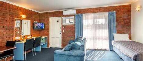 A hotel room with a brick wall, a TV, a desk with chairs, a sofa, and a bed.