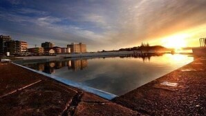 A waterfront swimming pool with buildings in the background. - Twenty Two North (Forster)