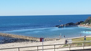 Piscina frente al mar con costa rocosa, señal de parada de autobús y vista despejada del océano.