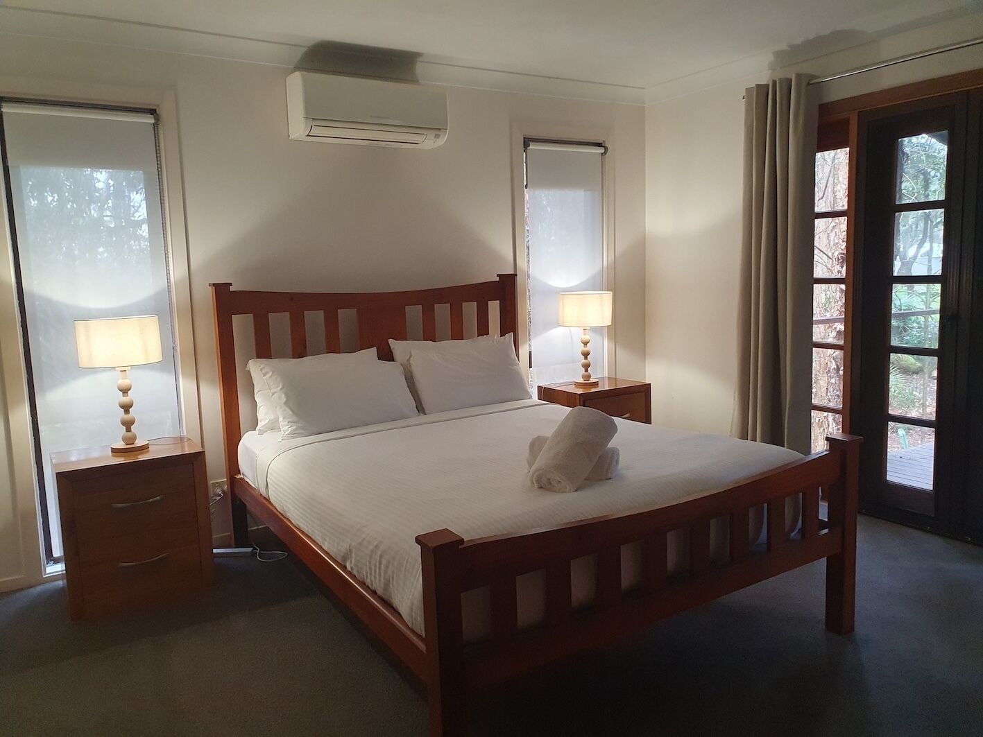 Una cama bien hecha con sábanas blancas, dos mesitas de noche con lámparas, un cabecero de madera y un ventanal con cortinas.
