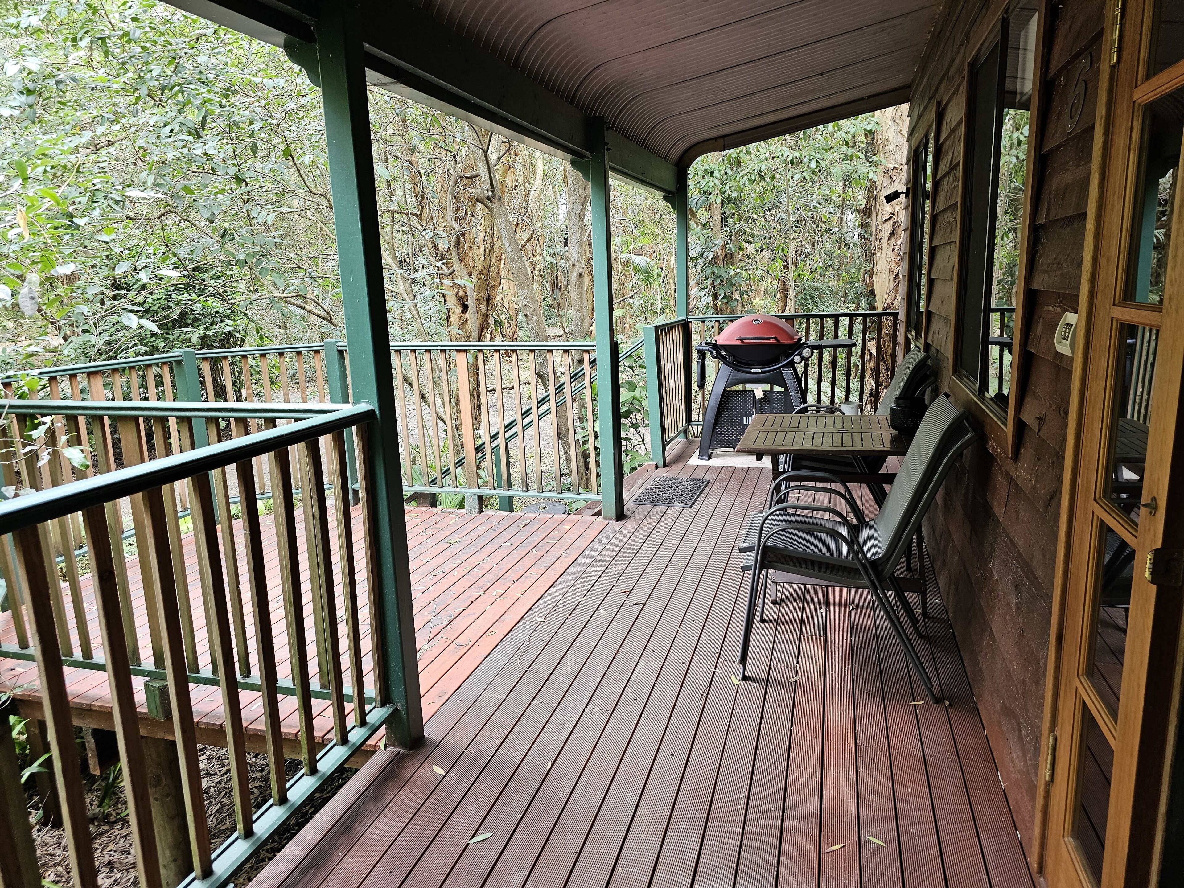 A wooden deck with a grill and chairs, surrounded by a green railing and trees.