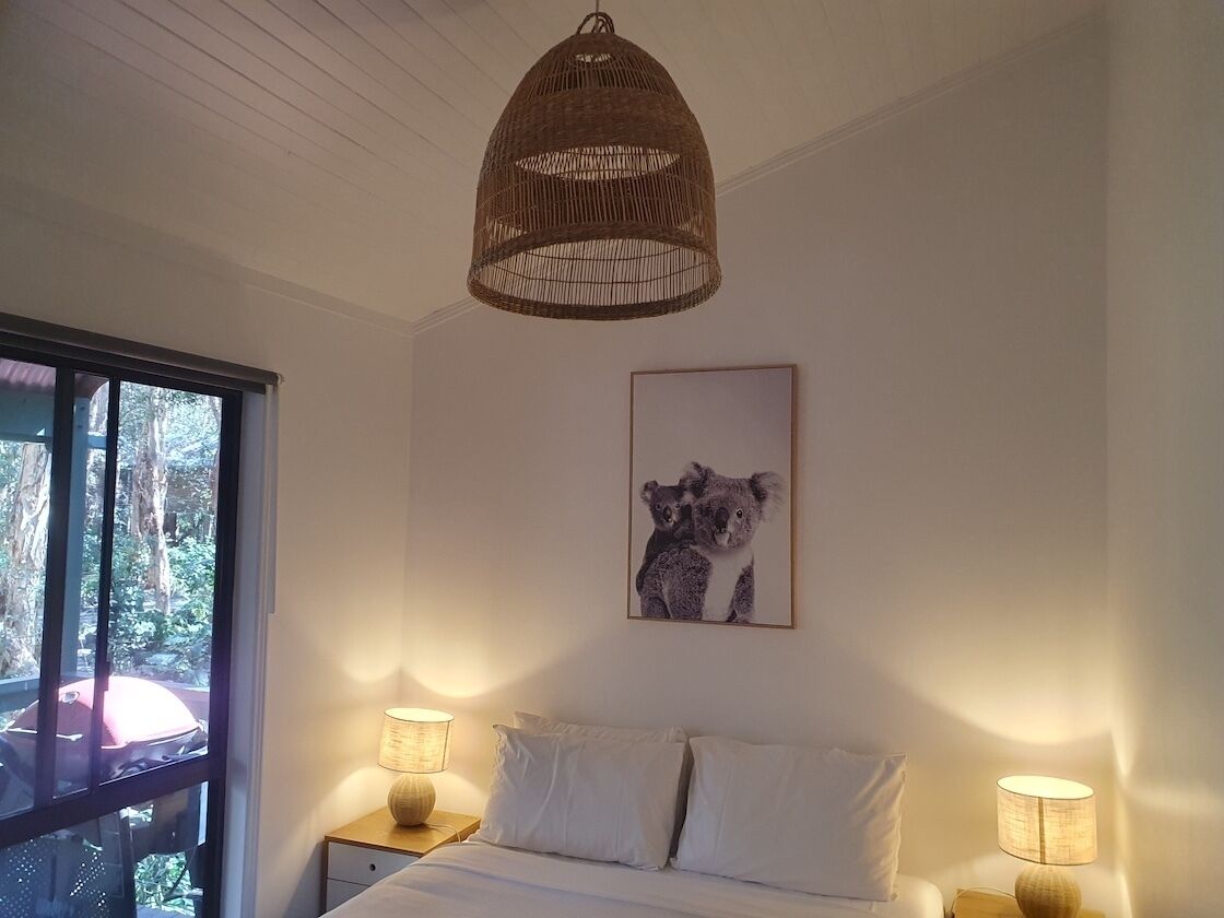 A bedroom with a bed, two bedside lamps, a framed picture of koalas, and a woven ceiling light fixture.