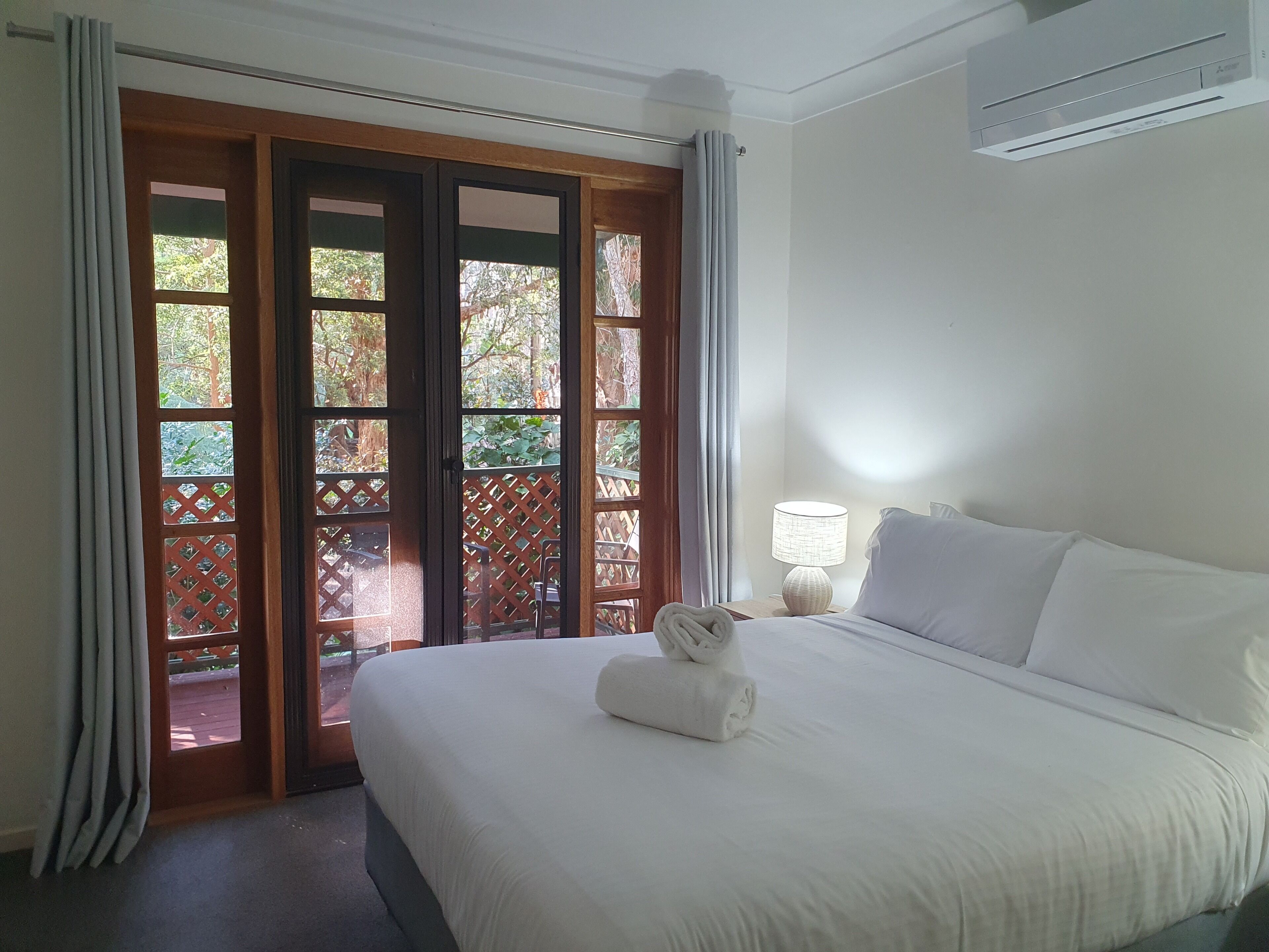 Une chambre d’hôtel avec un lit, une table de chevet avec une lampe et une vue sur l’extérieur par une porte en bois.