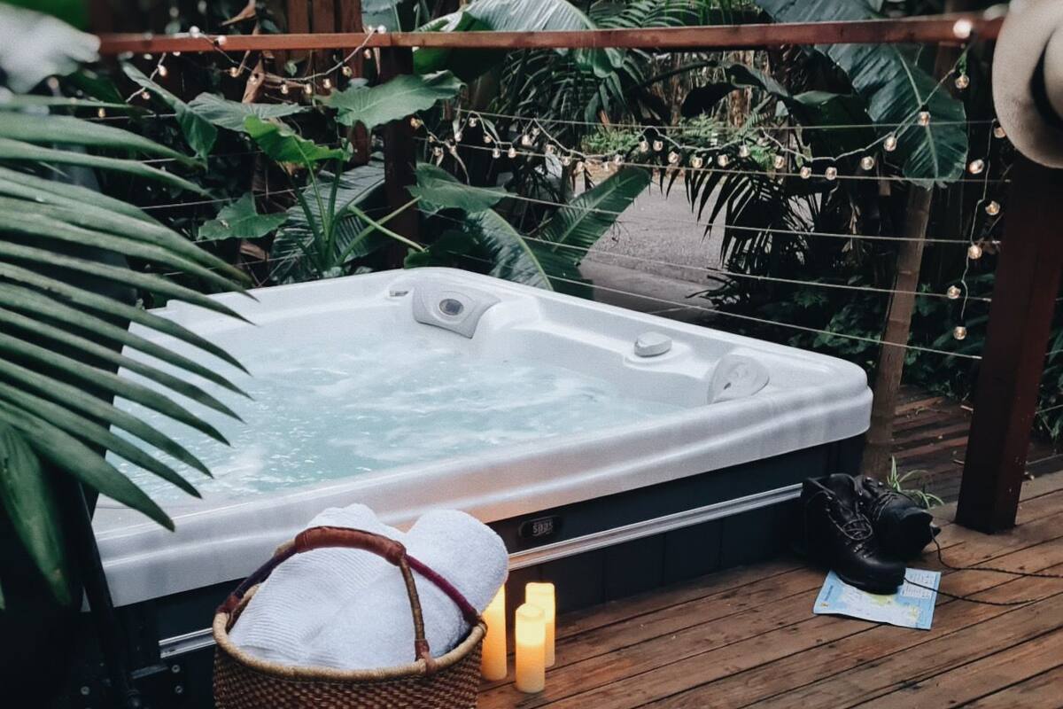 A hot tub surrounded by lush greenery, with a wooden deck and a basket of towels.