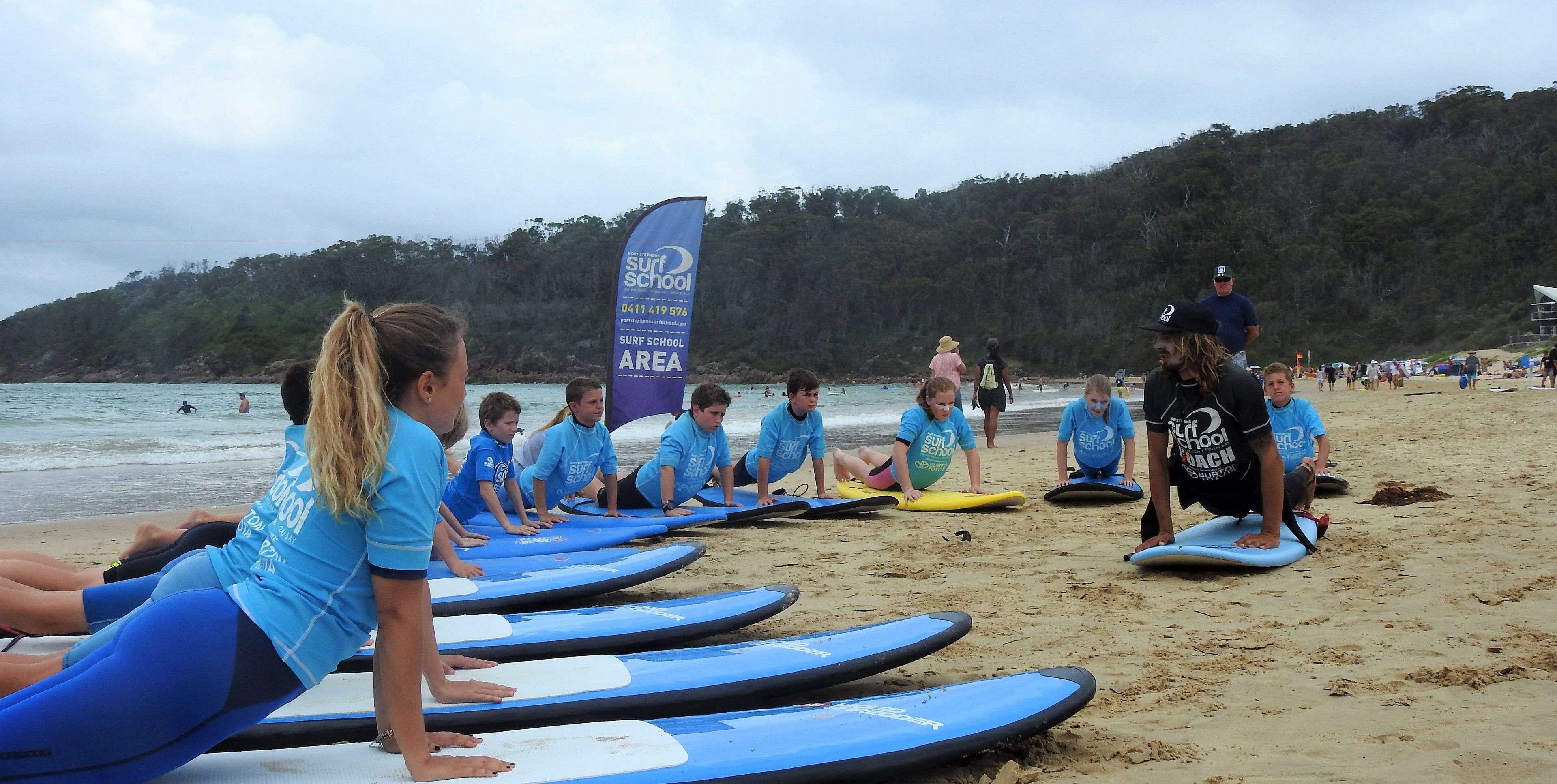 A group of people on a beach with surfboards, a blue banner, and a forested hill in the background.