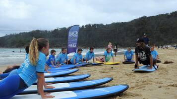 A group of people on a beach with surfboards, a blue banner, and a forested hill in the background.