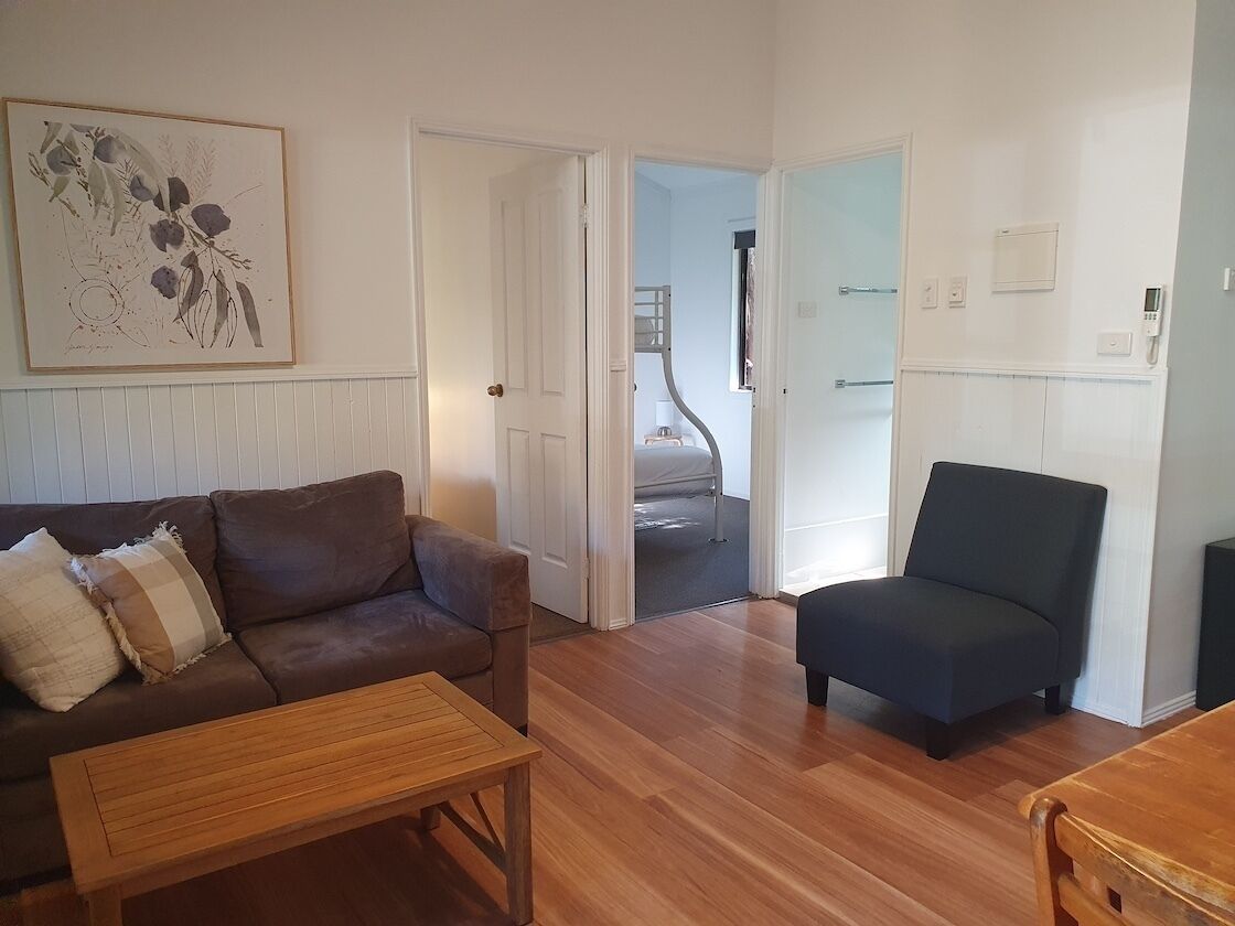 Sala de estar com um sofá marrom, mesa de centro de madeira e poltrona escura. Há uma porta que dá para um quarto com beliche.