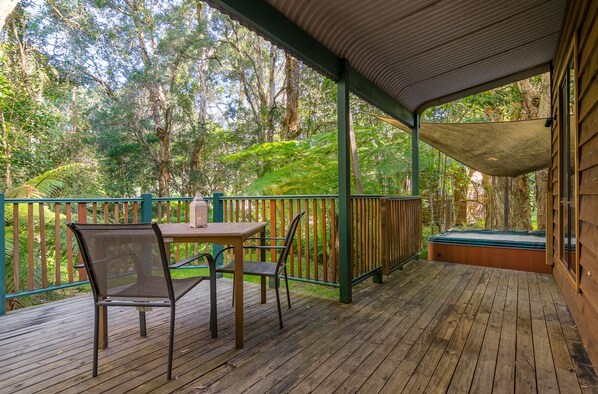 Una terraza de madera con mesa y sillas, rodeada por una cerca verde y árboles.