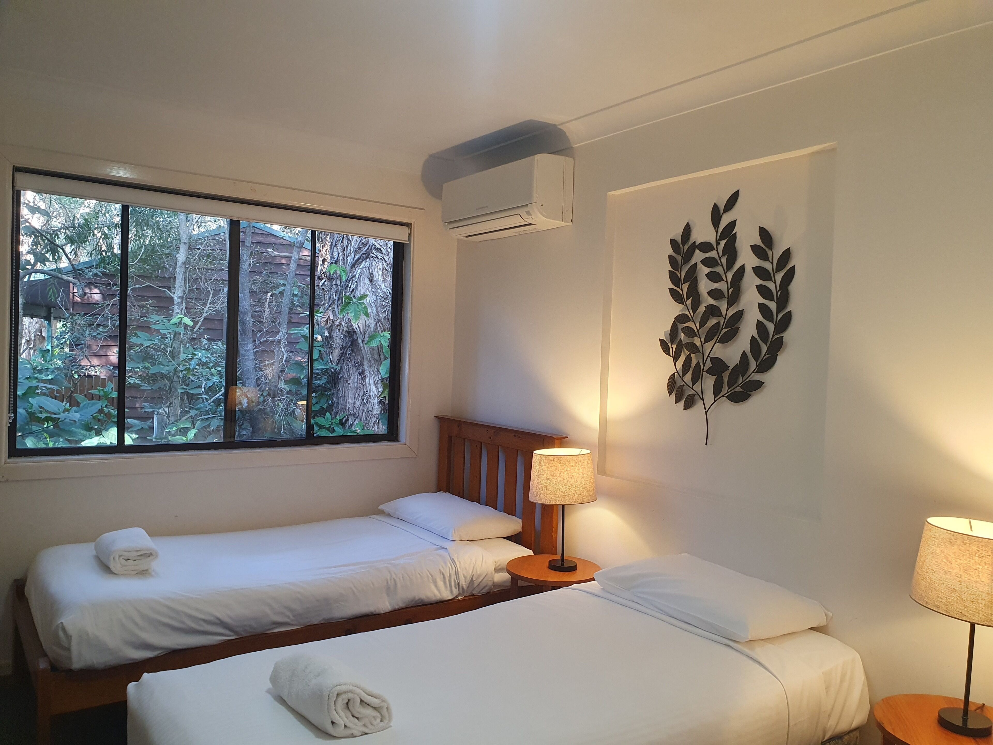 Habitación de hotel con dos camas, una ventana con vistas a árboles, aire acondicionado y una hoja decorativa en la pared.