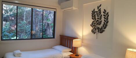 Habitación de hotel con dos camas, una ventana con vistas a árboles, aire acondicionado y una hoja decorativa en la pared.