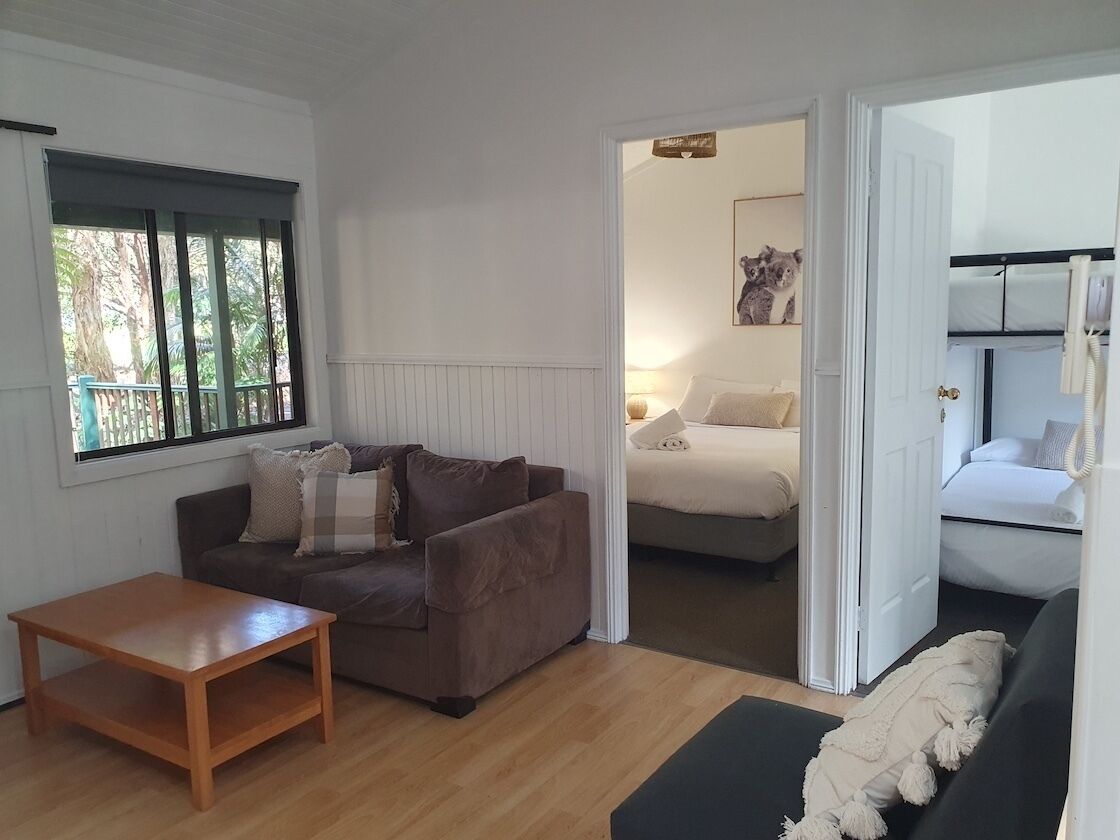 A cozy living room with a brown sofa, a wooden coffee table, and a black sofa. A bed with white bedding is visible through an open door.