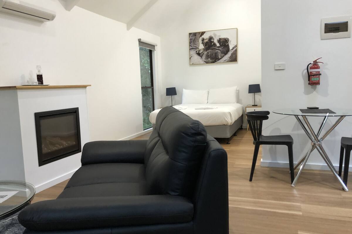 A modern living room with a black leather sofa, a glass coffee table, a fireplace, a bed with white bedding, and a small dining area with a glass table and two chairs.