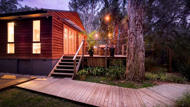 A wooden cabin with a deck, stairs, and outdoor seating area surrounded by trees. - Wanderers Retreat Port Stephens (Boat Harbour)
