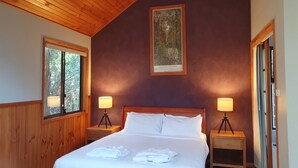 A bedroom with a wooden ceiling, a large bed with white linens, two bedside lamps, and a framed picture on the wall. - Wanderers Retreat Port Stephens (Boat Harbour)