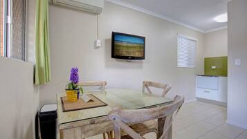 A dining area with a glass table, wooden chairs, a small potted plant, and a television mounted on the wall.