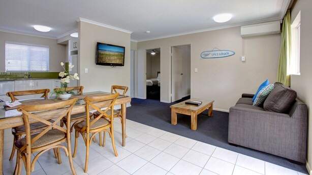 A modern living room with a dining area, a gray sofa, and a television on the wall. - Beachpark Apartments (Coffs Harbour)