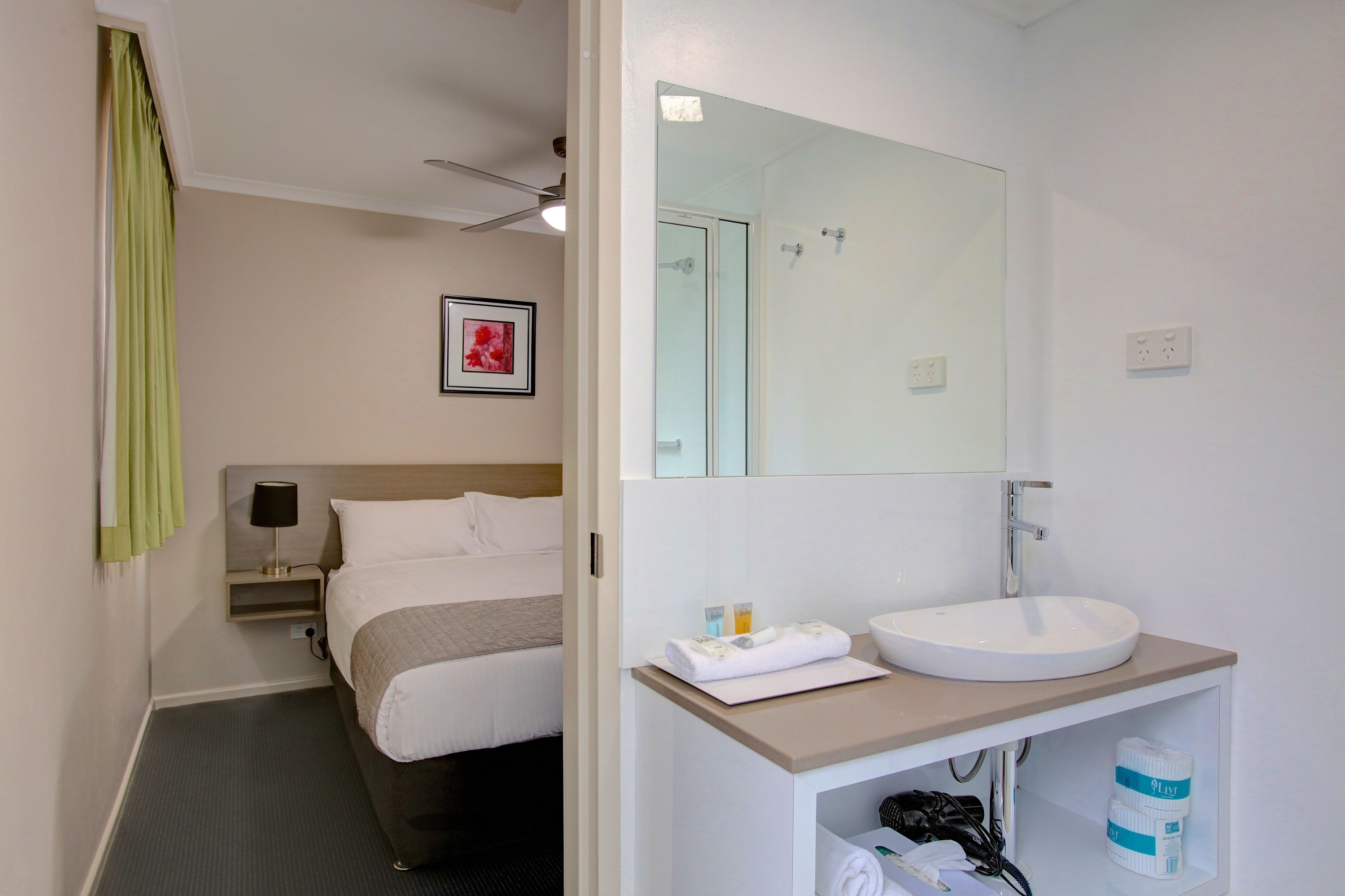 Une chambre d’hôtel comprenant un lit, une table de chevet avec une lampe, un grand miroir, un lavabo et un porte-serviettes.