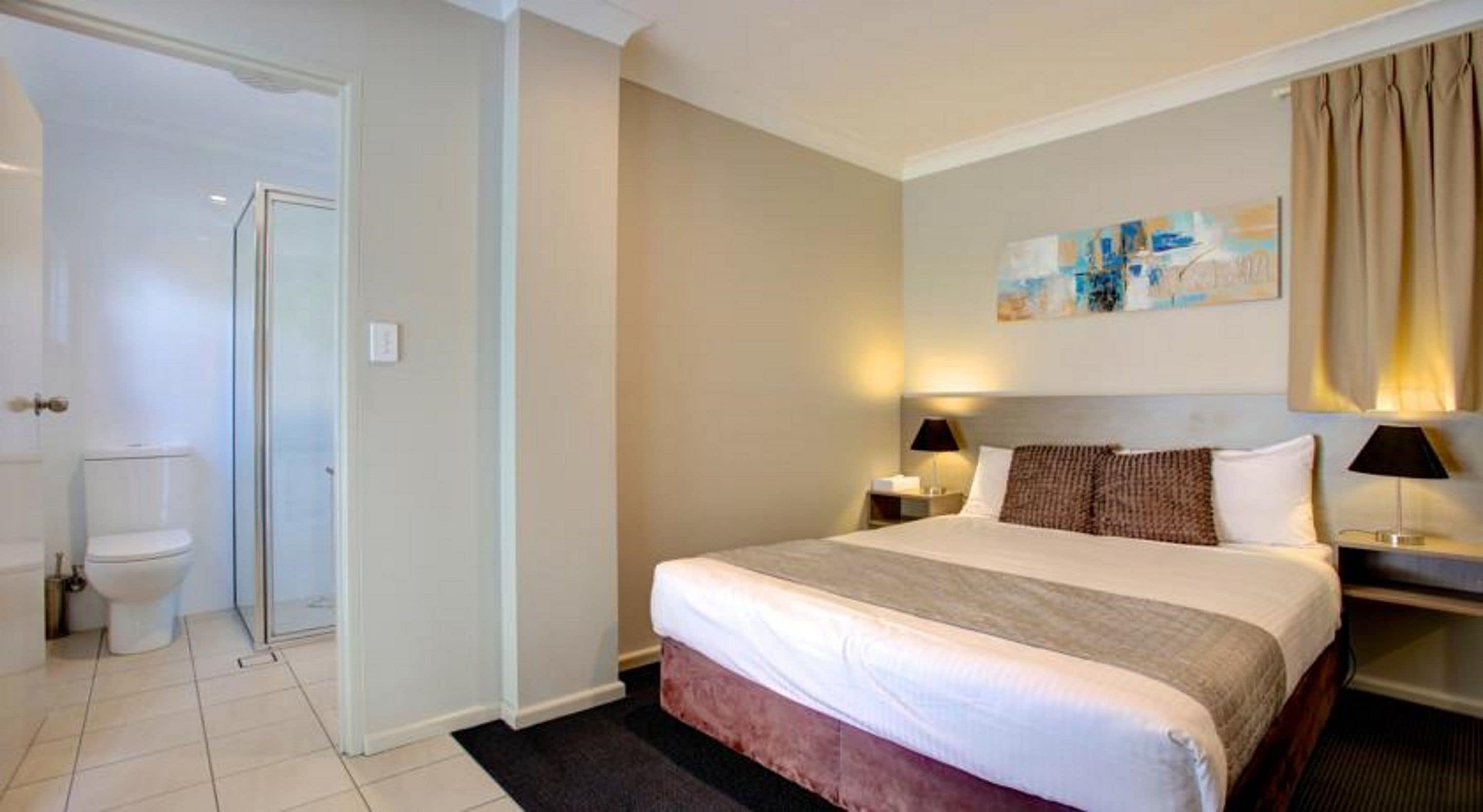 Une chambre d’hôtel comprenant un lit, deux tables de chevet, une commode, un tableau au mur et une salle de bain avec des toilettes et une douche.