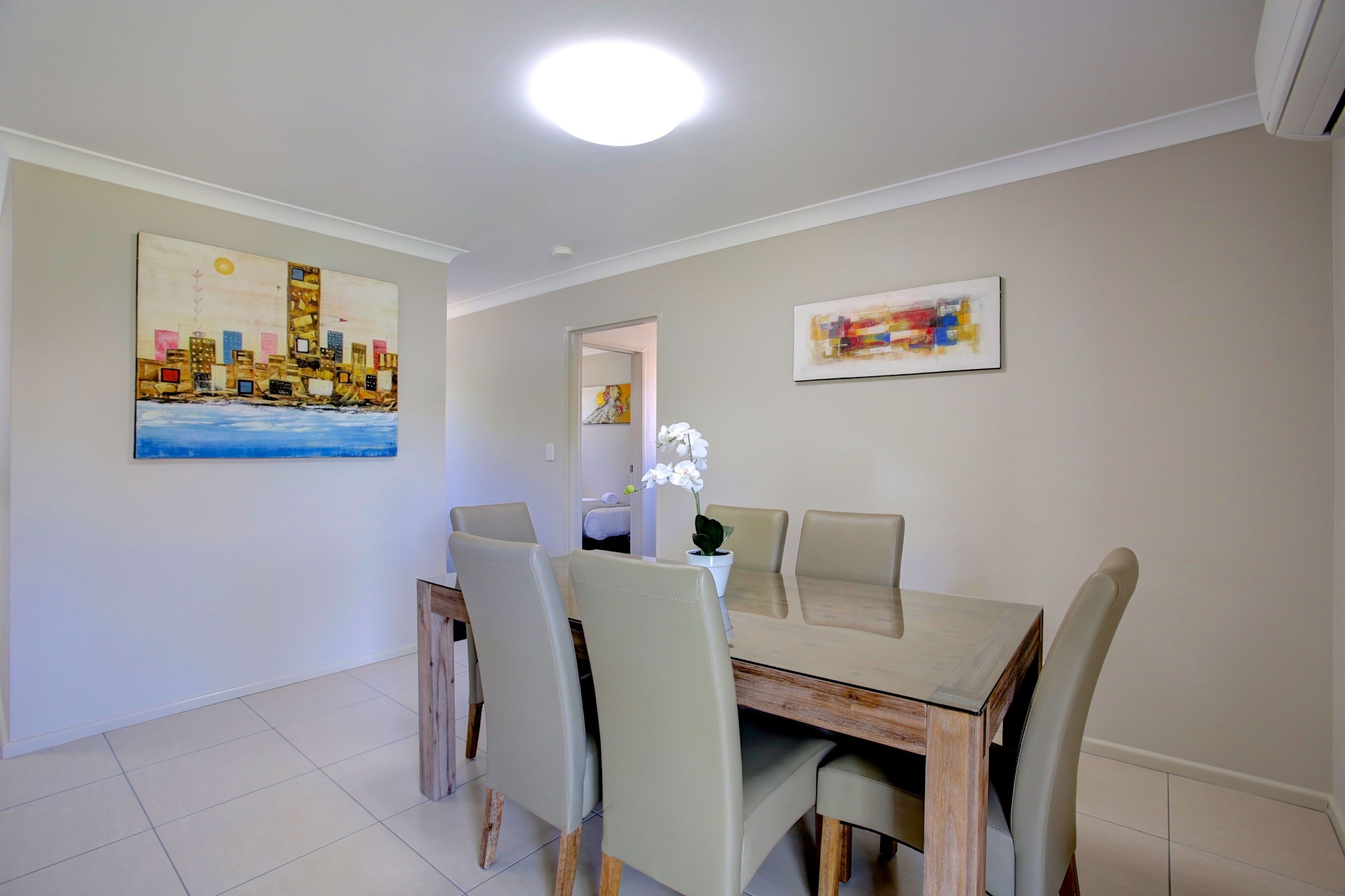 A dining area with a wooden table and four chairs, two paintings on the walls, and a ceiling light.