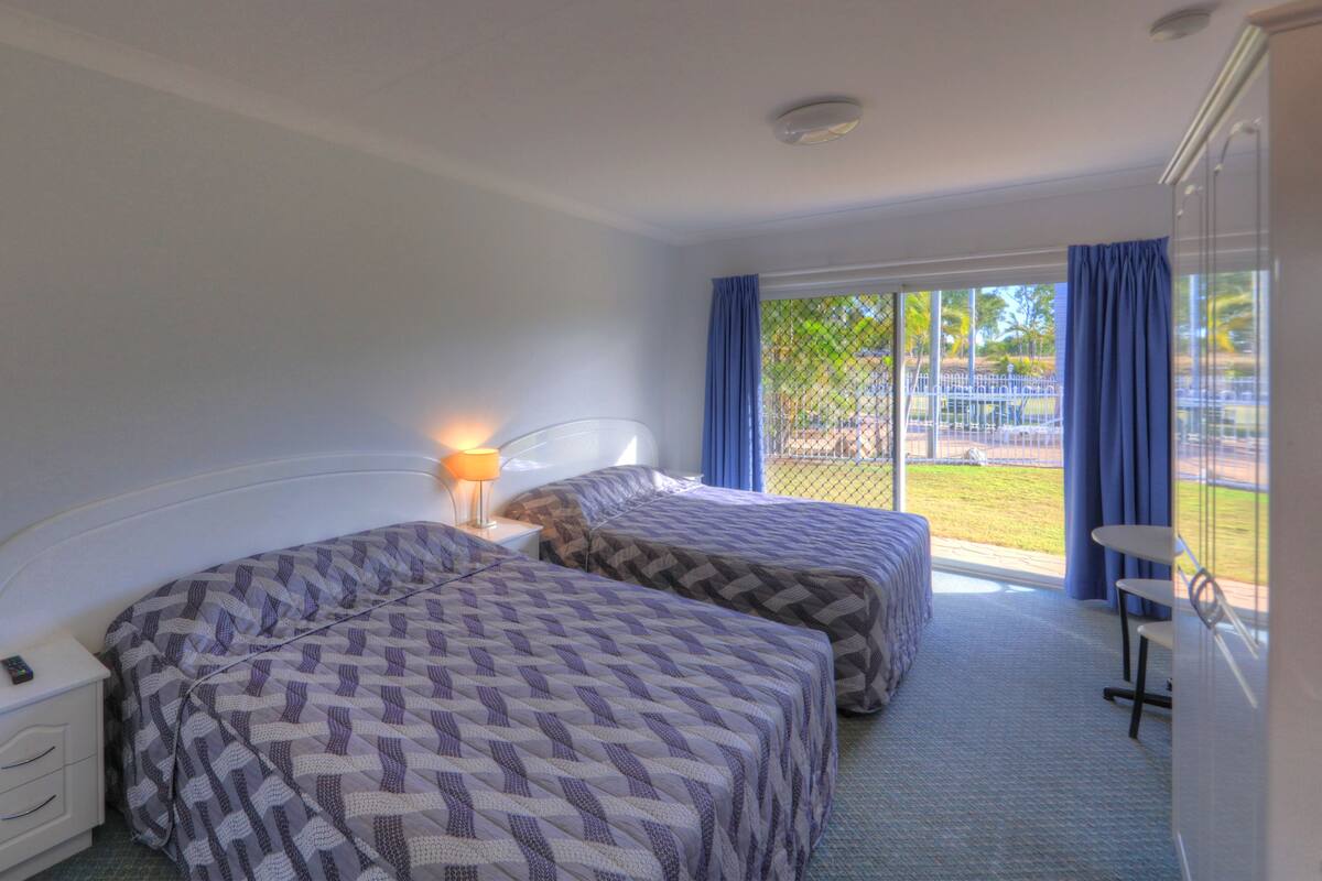 Habitación de hotel con dos camas, una mesita de noche, una lámpara y vistas a un espacio exterior.