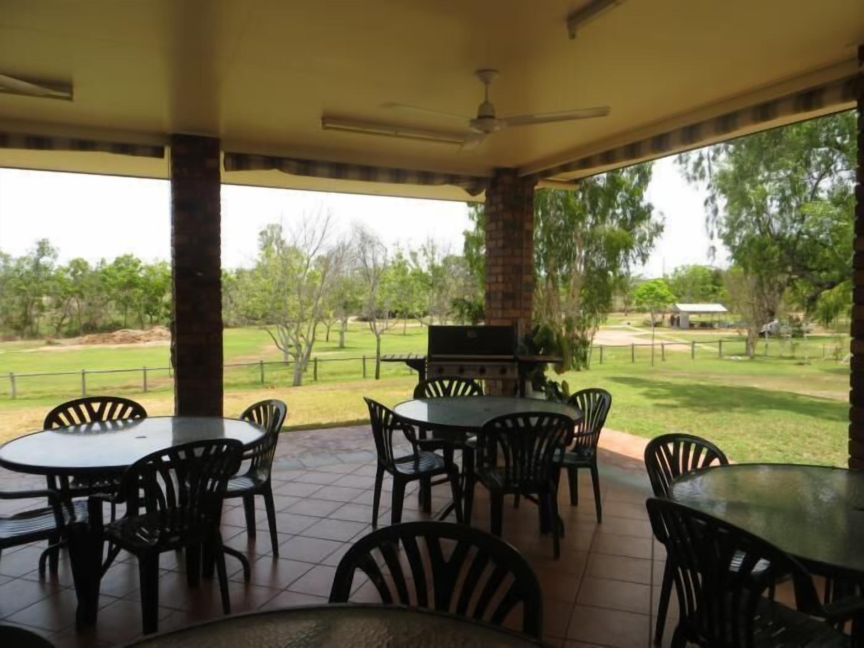 Un patio al aire libre con varias mesas y sillas redondas, un ventilador de techo y vistas a un campo cubierto de césped y árboles.