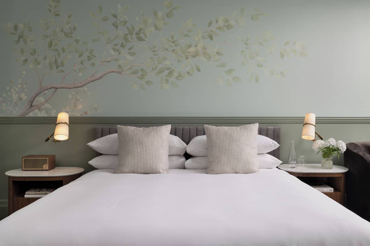 A neatly made bed with a white duvet, two bedside lamps, a small table with a vase of flowers, and a wall mural of a tree branch.