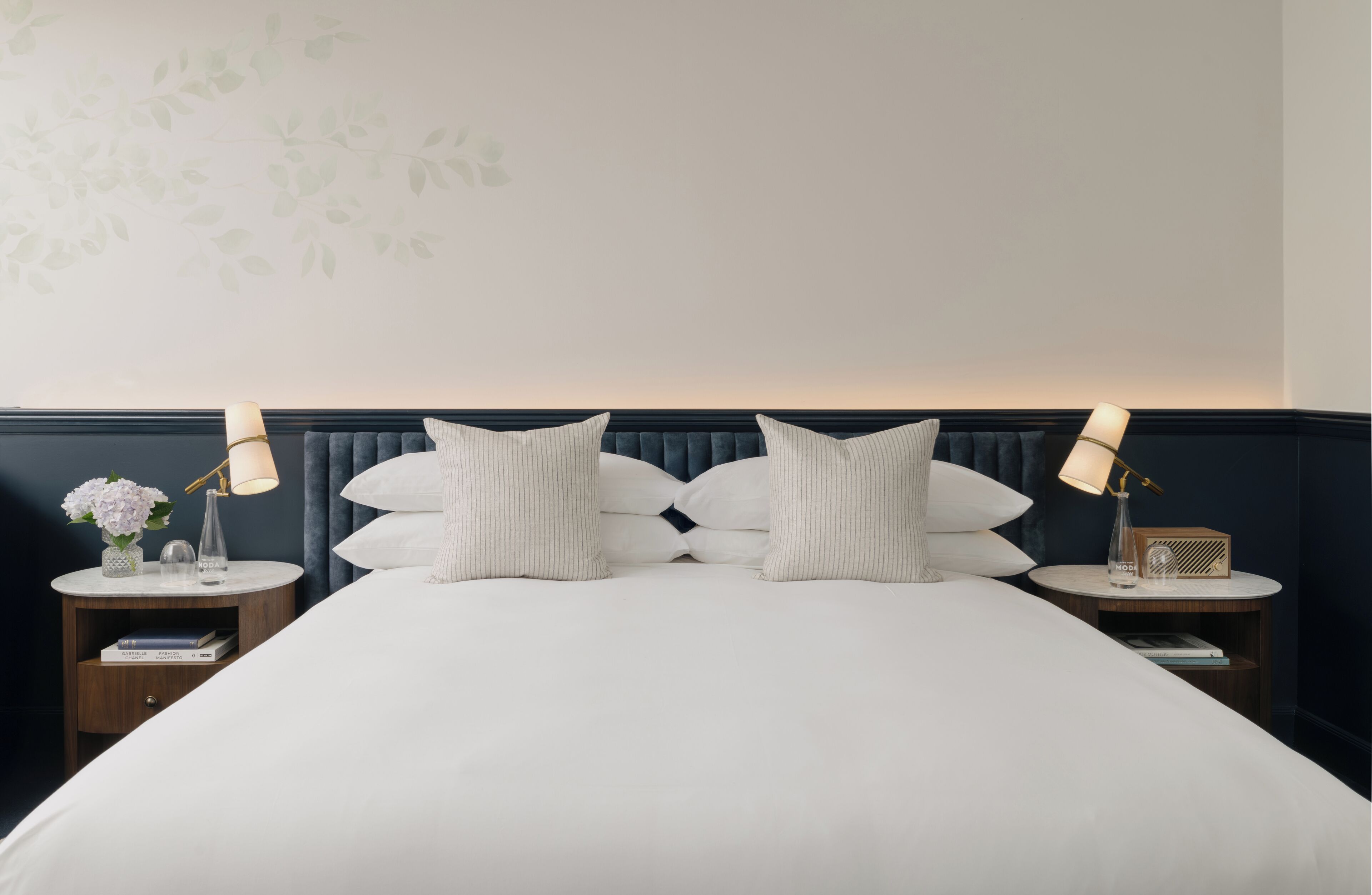 Una cama bien hecha con un cabecero oscuro, dos mesitas de noche con lámparas, y una pared con un diseño de hojas.