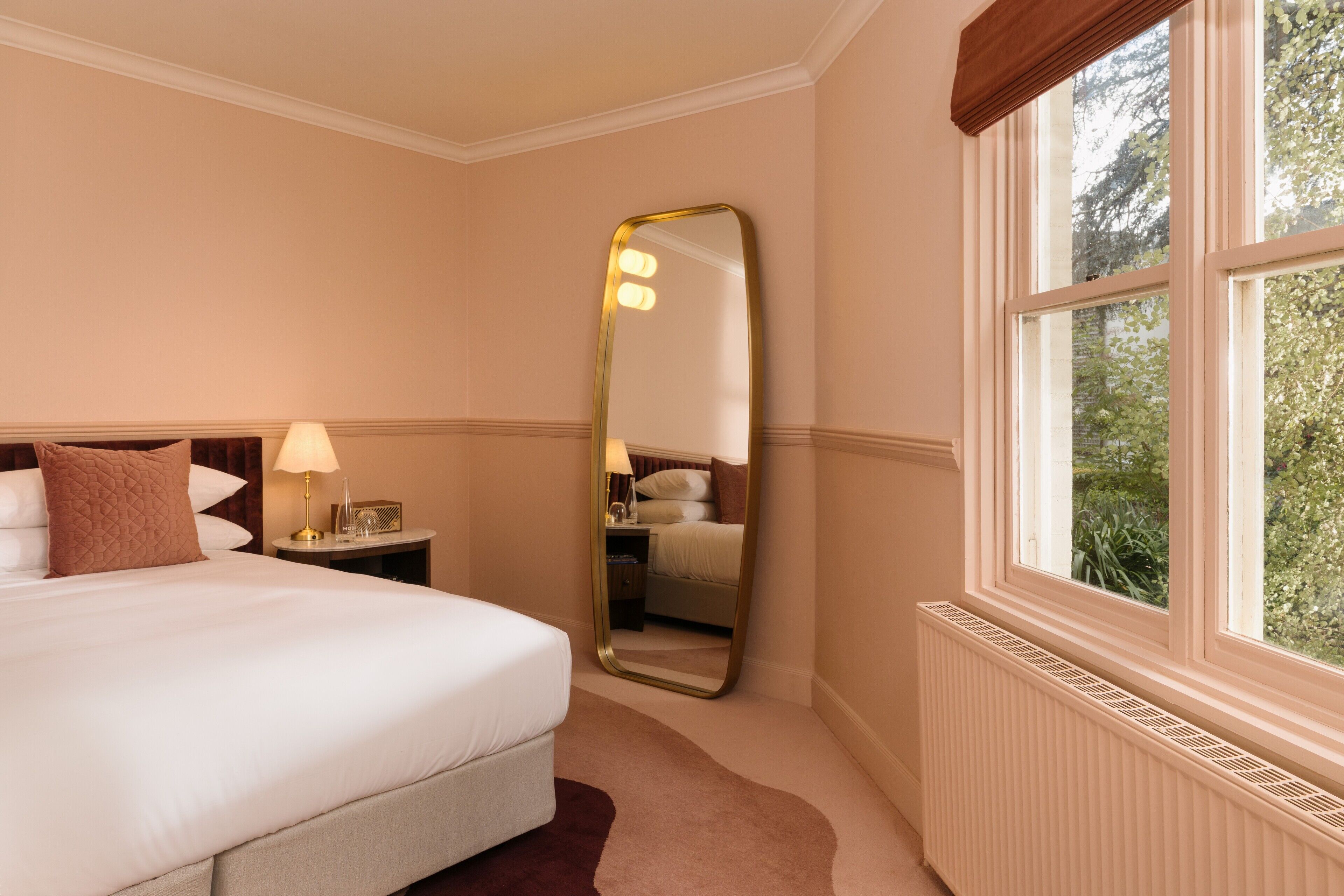 A hotel room with a large bed, a nightstand with a lamp, a mirror, and a window with a view of greenery.