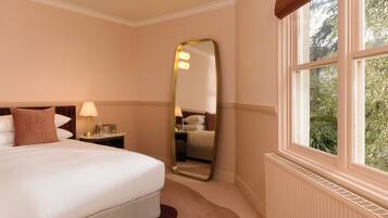 A hotel room with a large bed, a nightstand with a lamp, a mirror, and a window with a view of greenery.