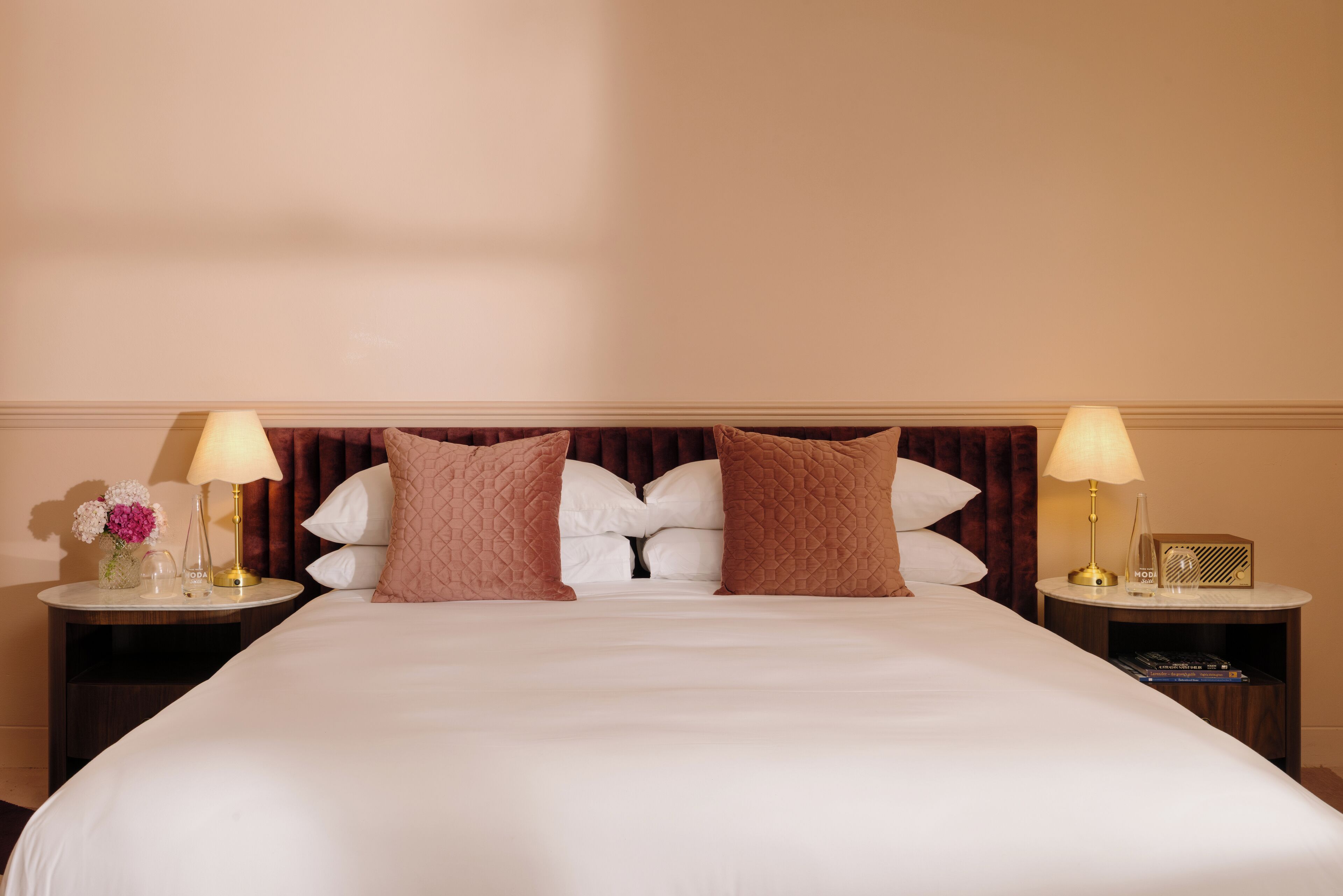 A hotel room with a large bed, two bedside tables, a nightstand, a lamp, a vase with flowers, and a radio.