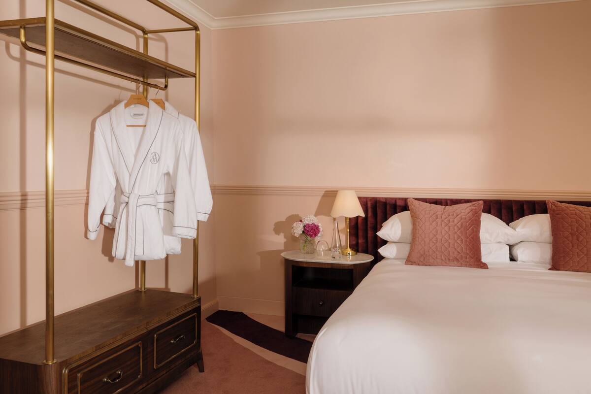 A hotel room with a bed, a bedside table with a lamp, a nightstand, a robe hanging on a rack, and a small vase with flowers.