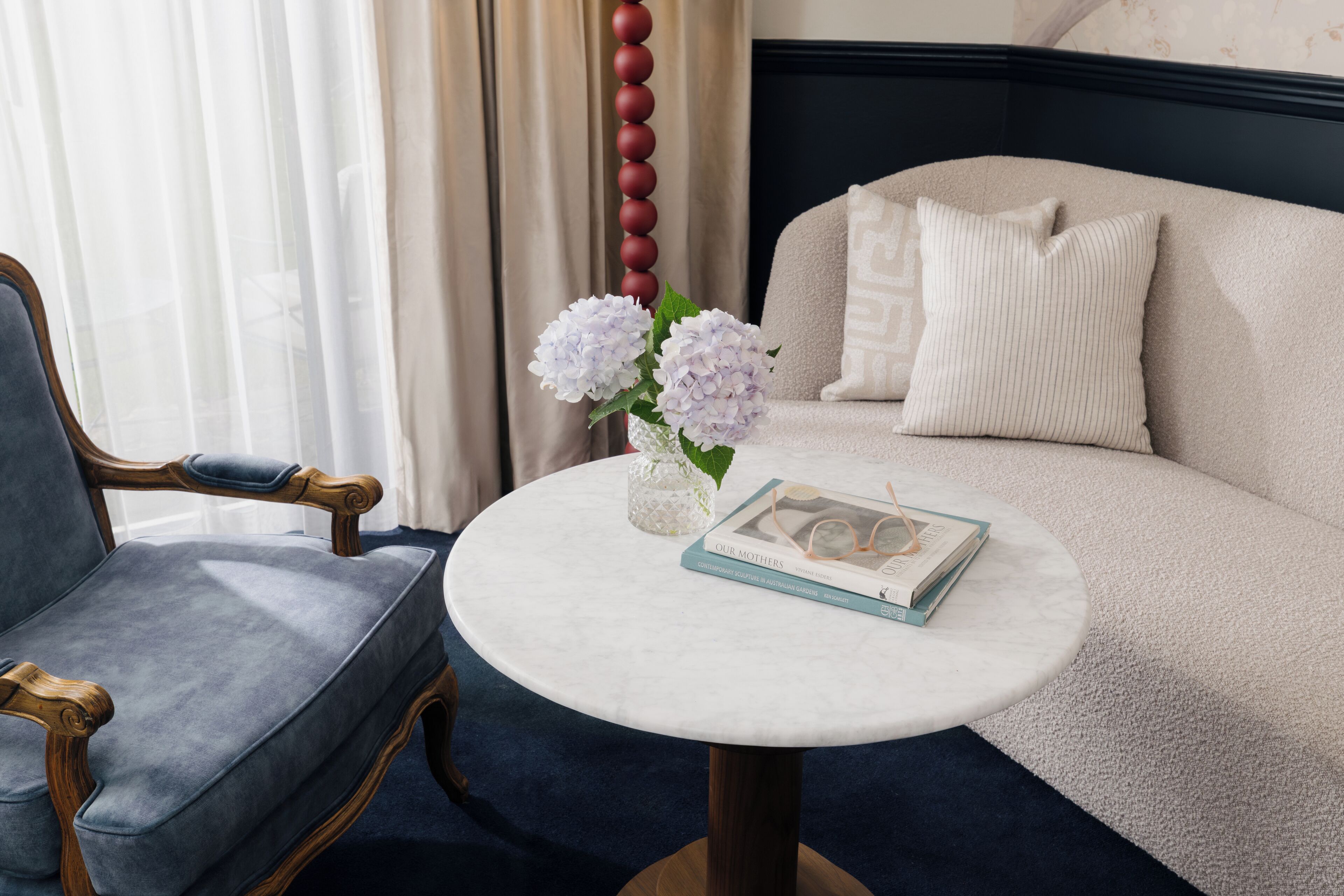 Une pièce avec une table ronde, un fauteuil bleu, un canapé beige et un vase rempli de fleurs.