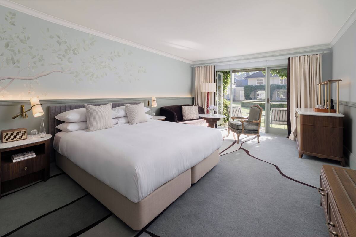 A spacious bedroom with a large bed, a side table, a chair, and a view of the outdoors through a sliding door.