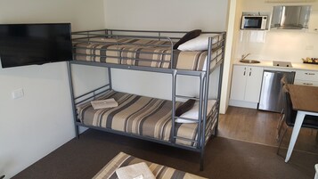 Un letto a castello con biancheria a righe, una TV a schermo piatto montata a parete, un angolo cottura con forno a microonde ed elettrodomestici in acciaio inossidabile, e un piccolo tavolo di legno.