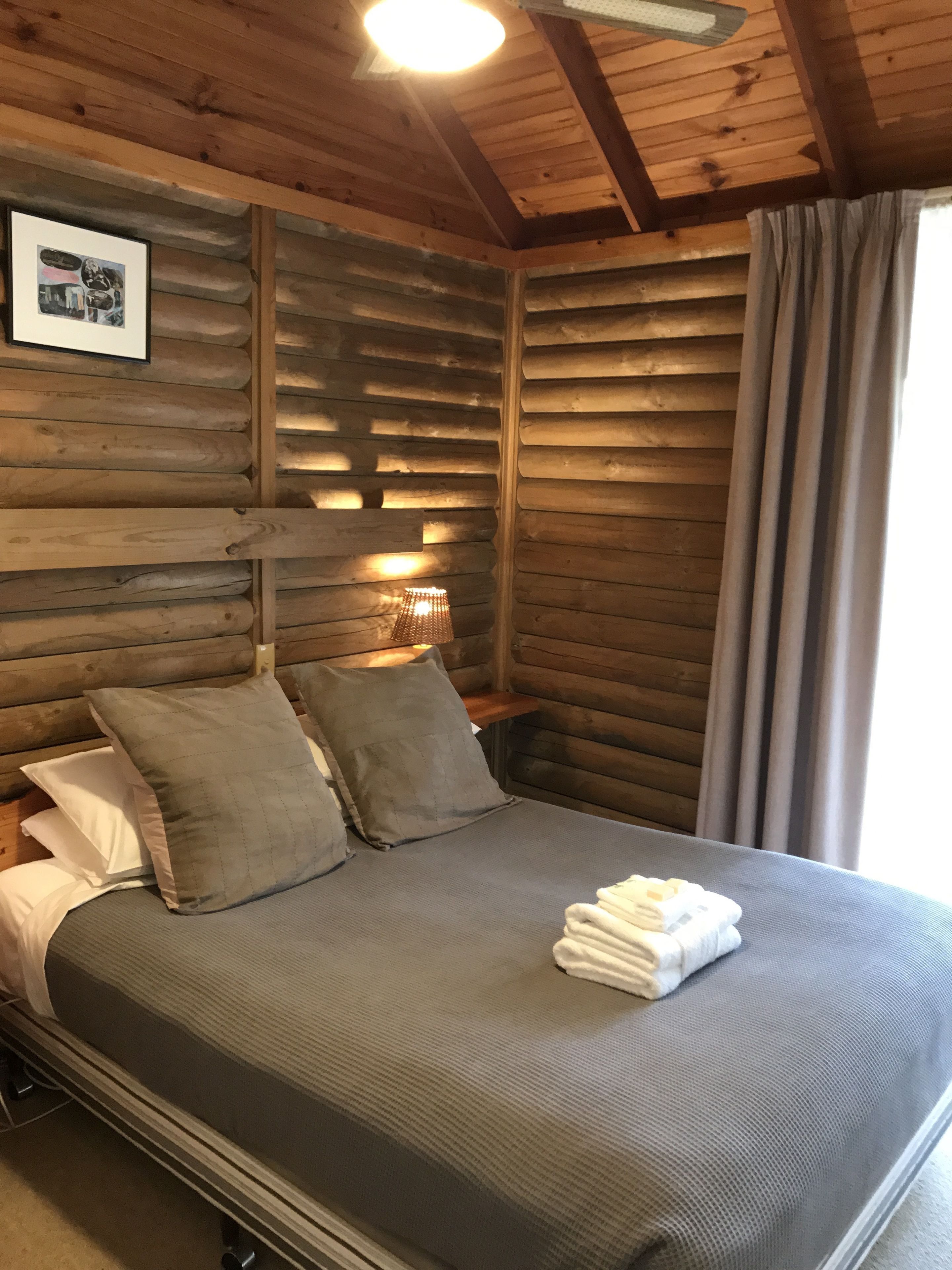 A wooden cabin room with a bed, pillows, a nightstand, and a framed picture on the wall.
