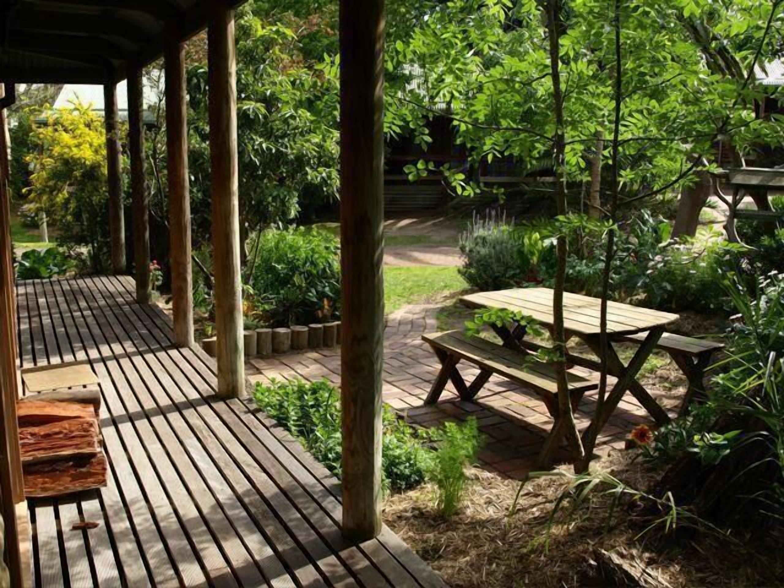 Una terraza de madera con una mesa de picnic y bancos, rodeada de abundante vegetación y un sendero de jardín.