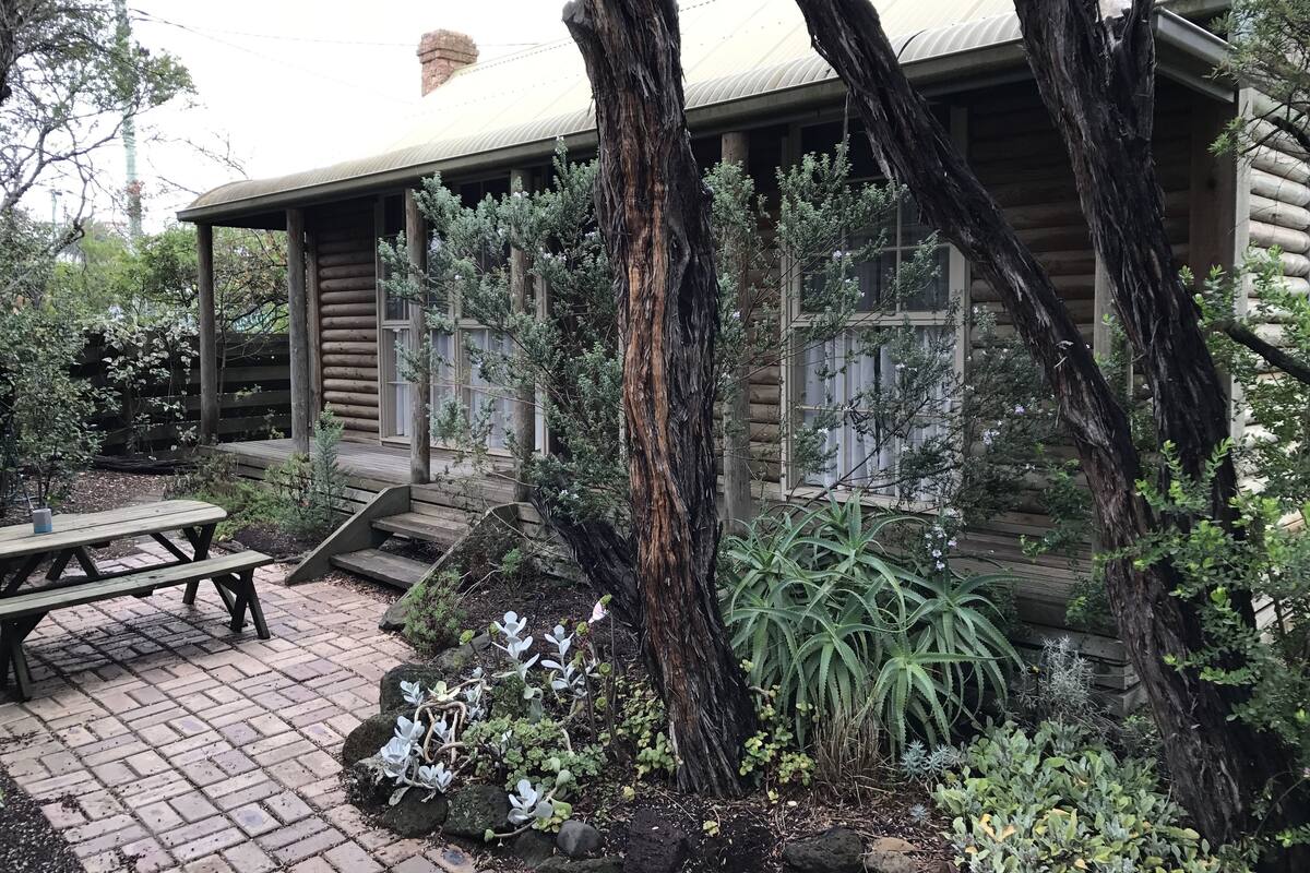 A rustic wooden cabin with a porch, surrounded by lush greenery and a brick patio.