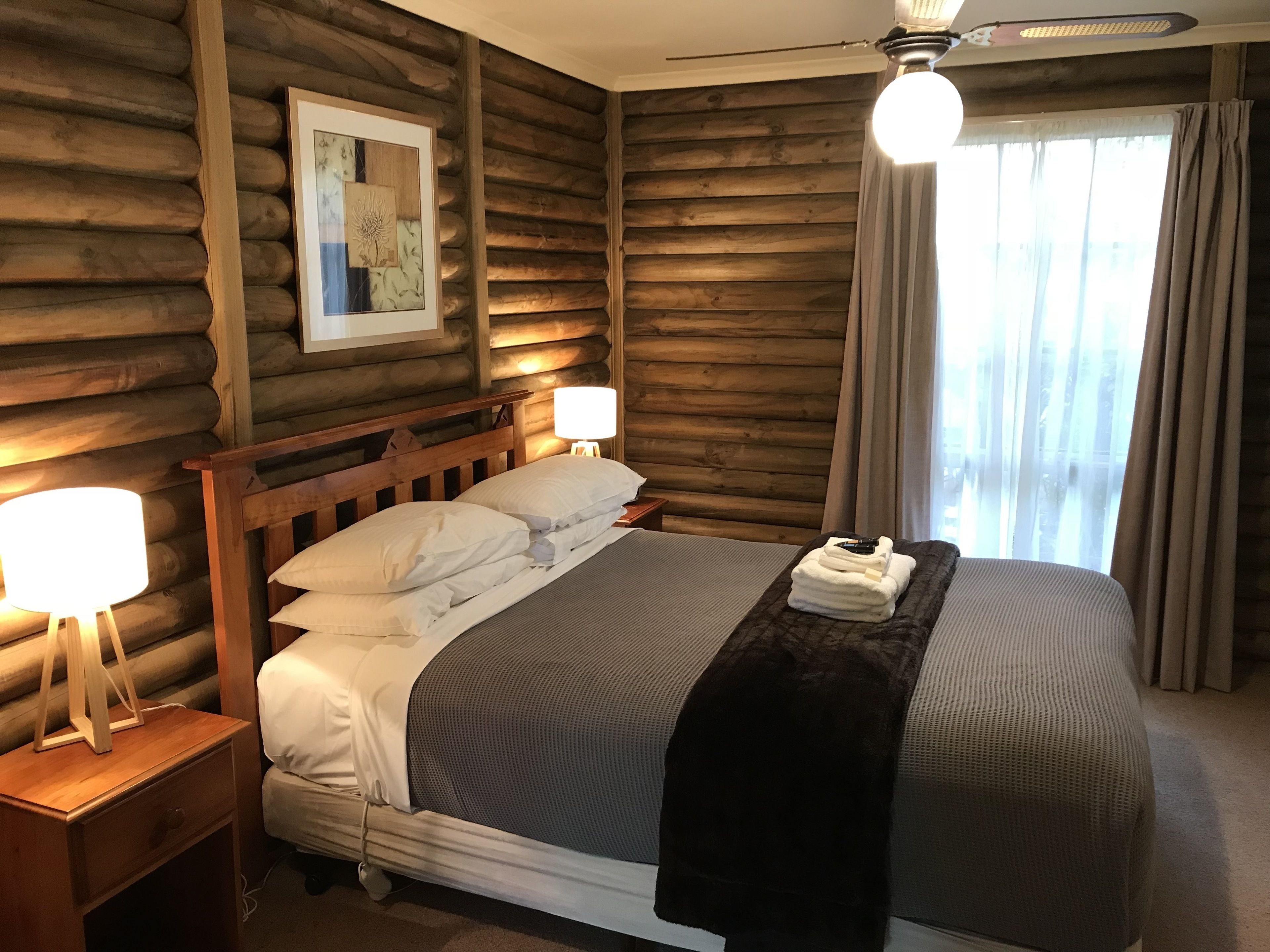 A wooden log cabin-style bedroom with a bed, two bedside lamps, a framed picture on the wall, and a window with curtains.