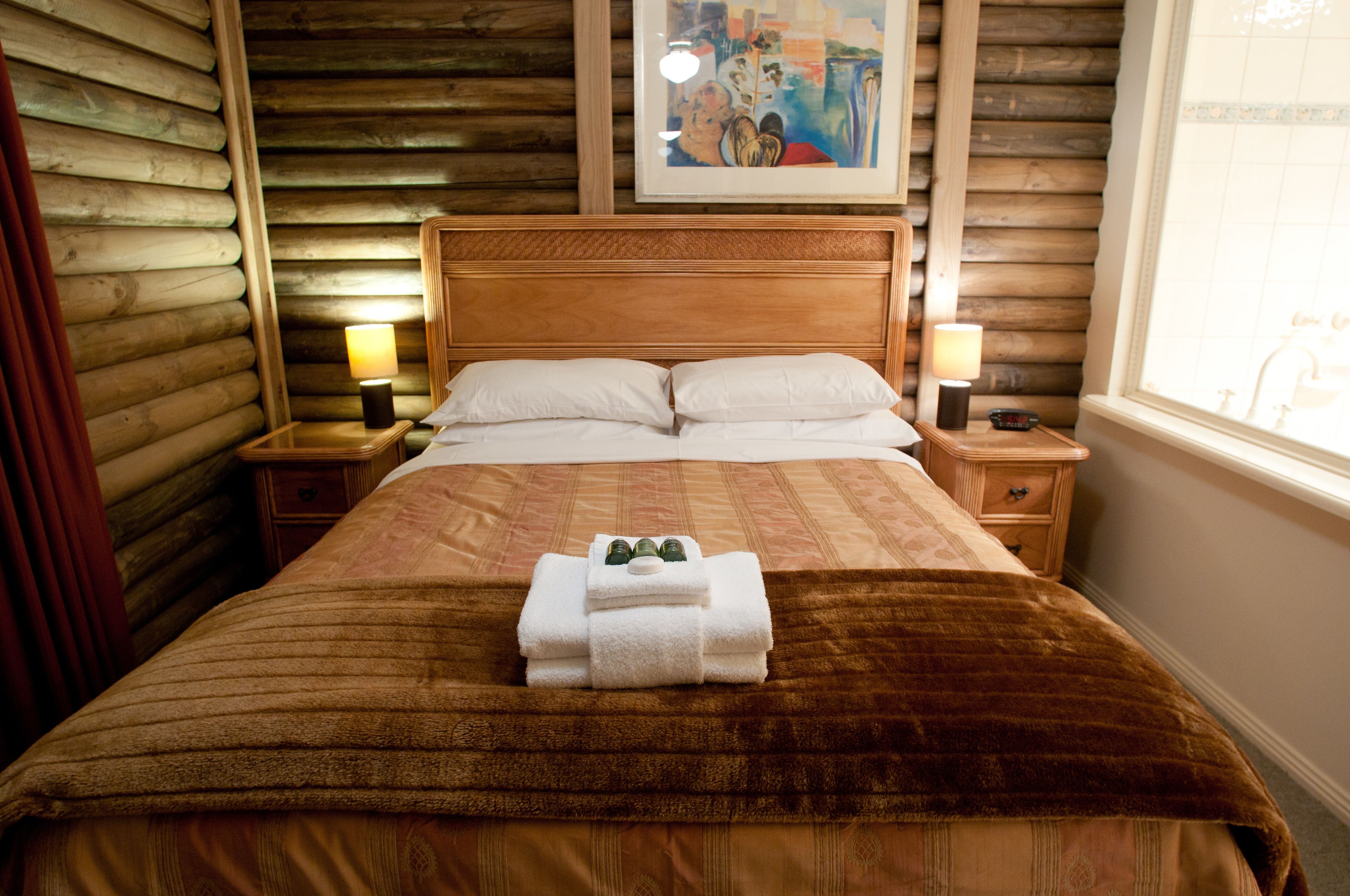 A neatly made bed with a brown blanket and white pillows, a wooden headboard, two bedside lamps, and a framed artwork above the bed.