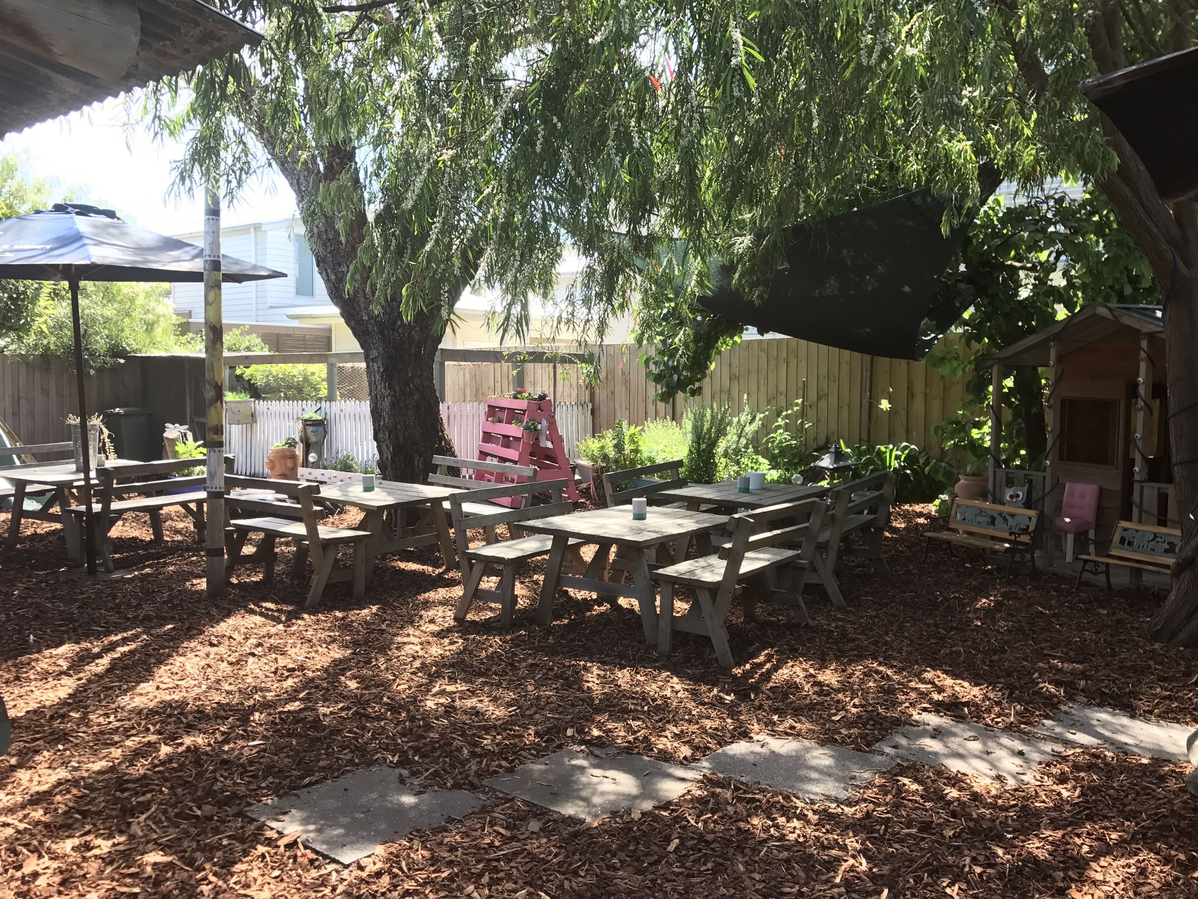 Un patio al aire libre con mesas y bancos de madera, rodeado de árboles y una cerca de madera.