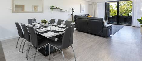 A modern dining area with a table set for six, a black leather sofa, and a balcony with a view of trees.
