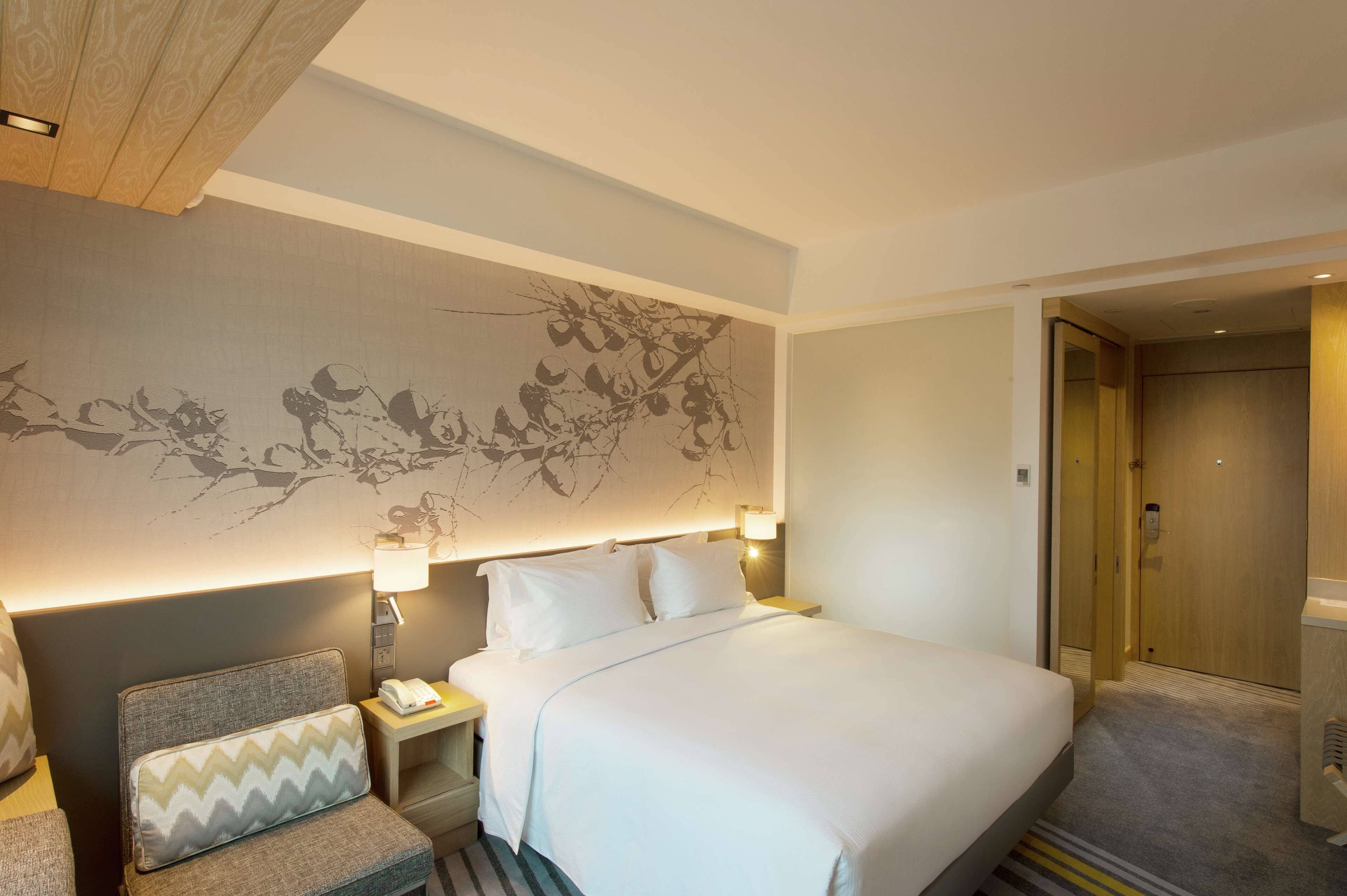 Quarto de hotel com uma cama grande, uma mesinha de cabeceira com abajur, uma cadeira com travesseiro e uma parede com um mural floral.