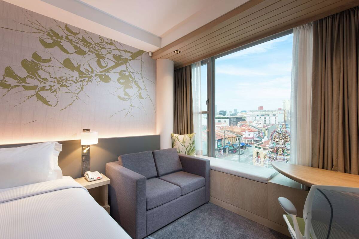 Habitación de hotel con cama, sofá gris, escritorio con silla, ventana con cortinas y vista a la ciudad.