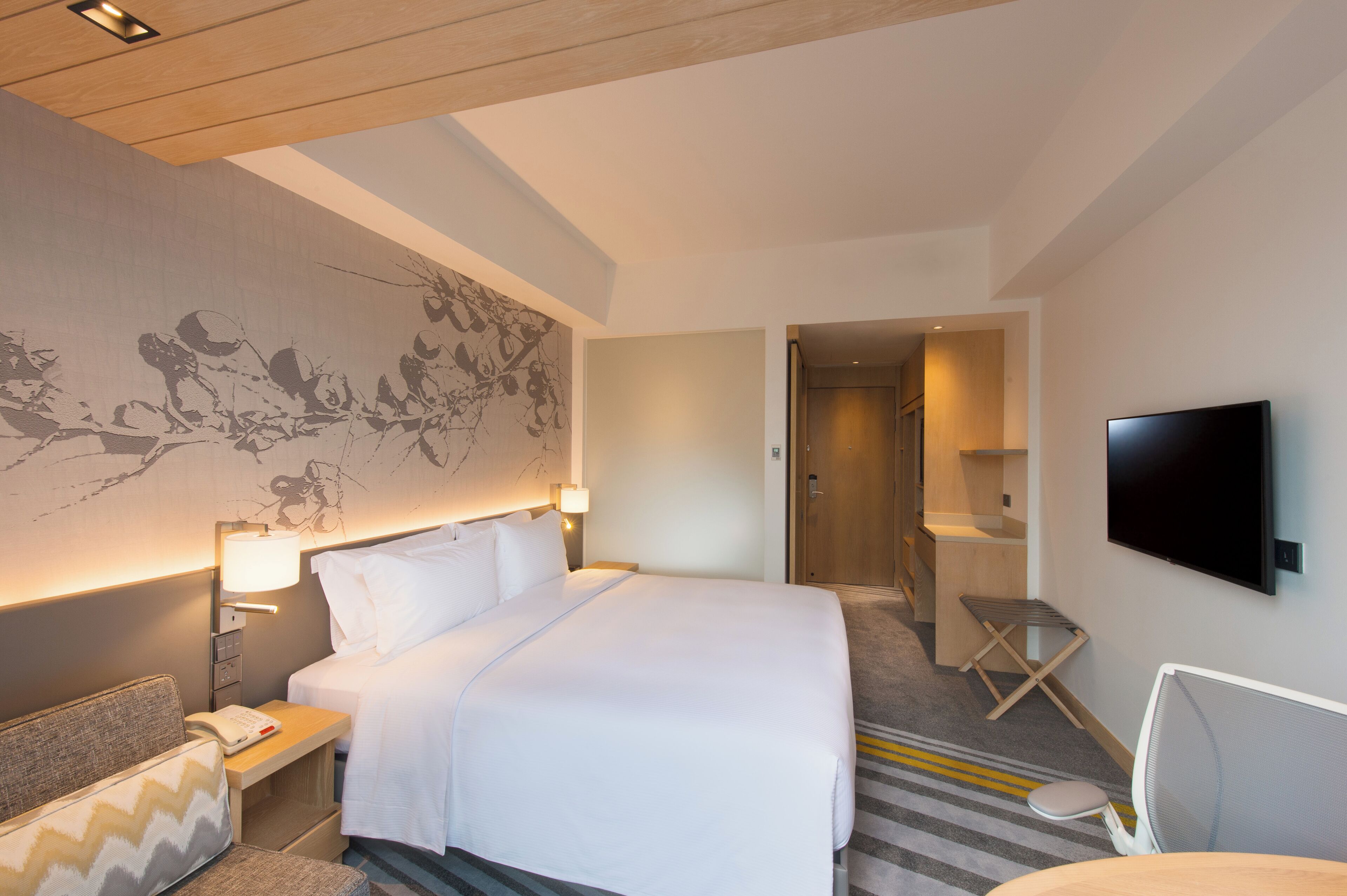 A hotel room with a large bed, bedside table with a lamp, a flat-screen TV mounted on the wall, and a small seating area with a chair and a bench.