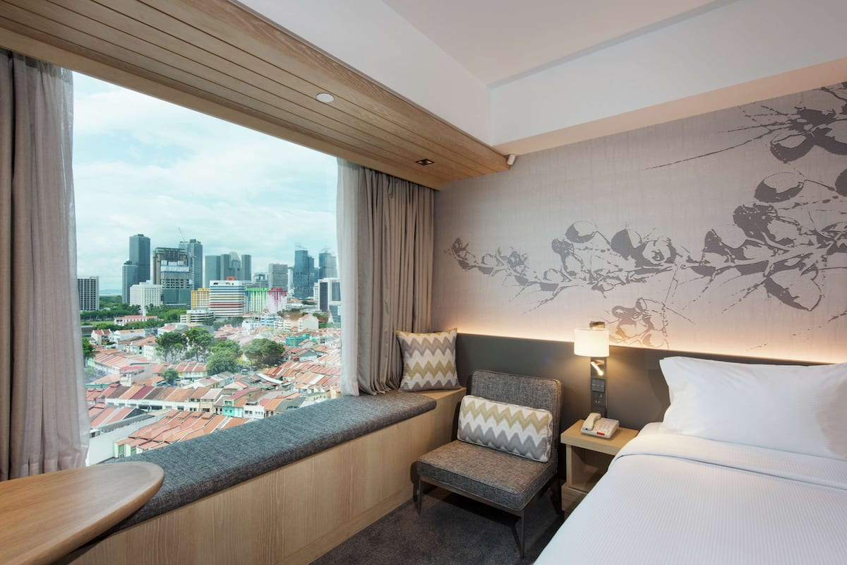 Habitación de hotel con vista a la ciudad, una cama, una silla, una mesita de noche con lámpara y un ventanal con cortinas.