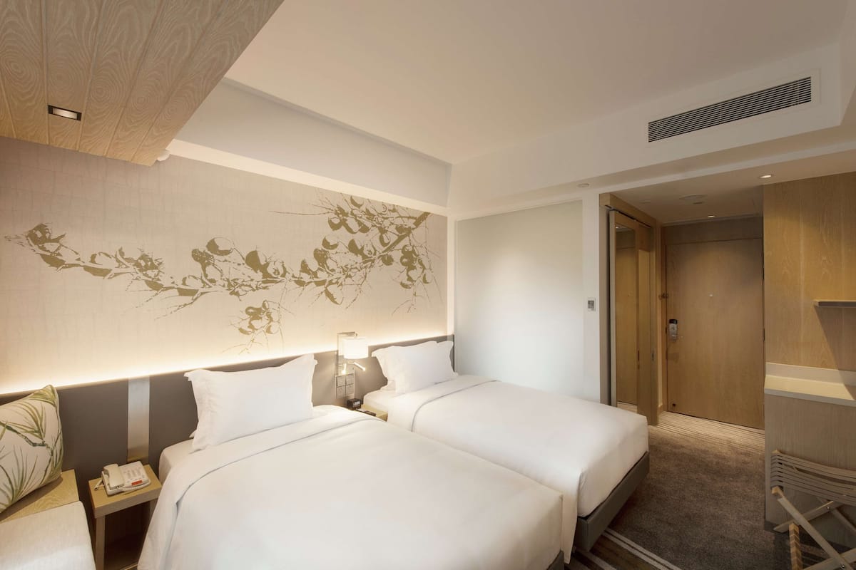 Una habitación de hotel con dos camas, una mesita de noche con teléfono, una lámpara de pared y una puerta que da a otra habitación.