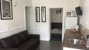 Un espacio habitable compacto con un sofá marrón oscuro, una pequeña kitchenette con fregadero y una cama al fondo.
