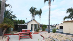 Zona residencial con una casa, un patio con banca roja y silla blanca, palmeras y un camino de entrada de grava.